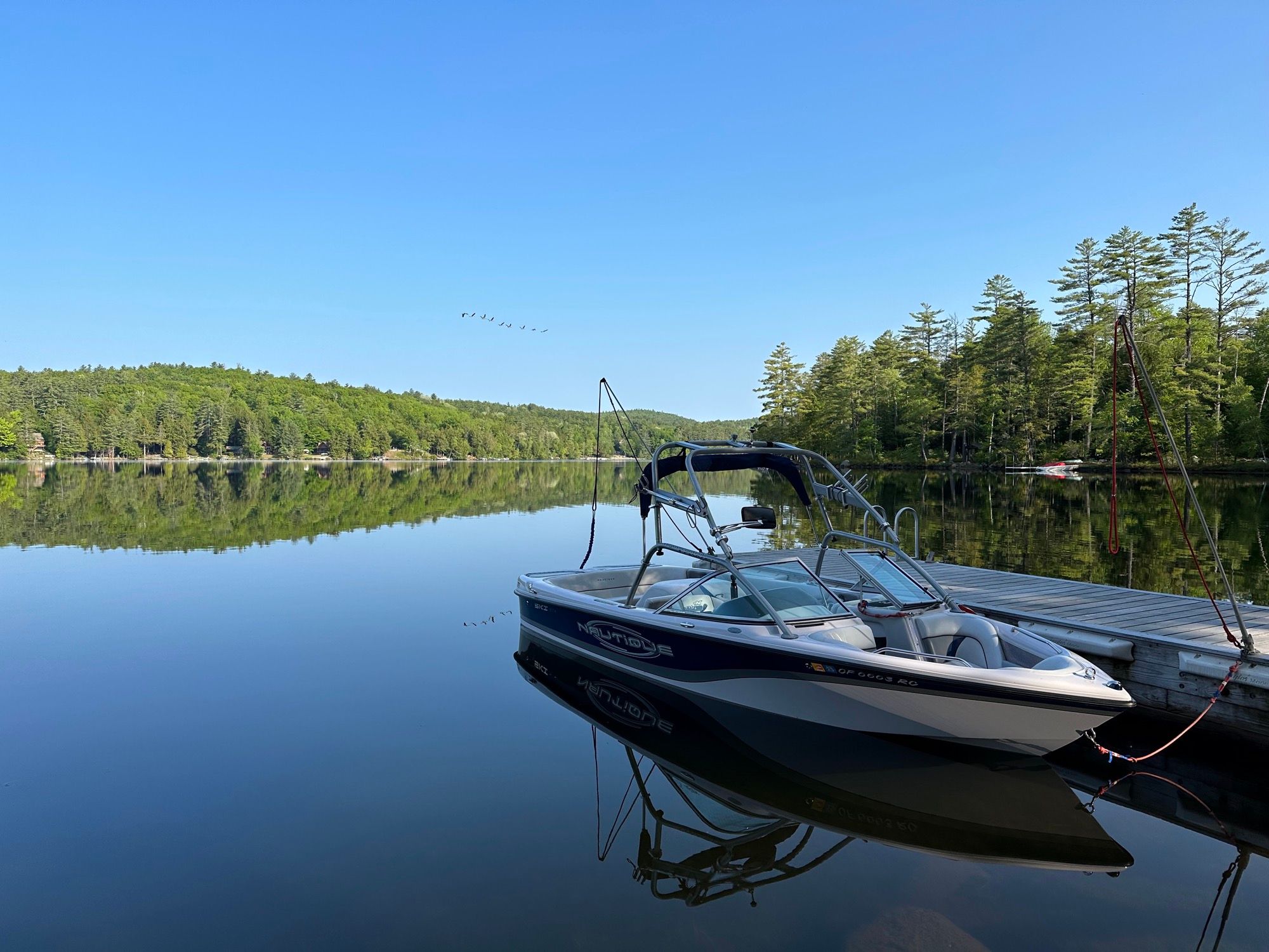A ski boat tied to the dock at Camp Sunnyside on a glassy morning, geese overhead.